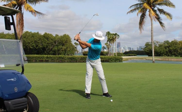 A person in a blue shirt and white pants at the start of their golf swing.